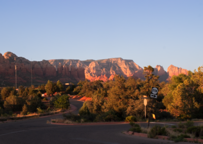 A scenic view of a towering red rock mountain in Sedona with jagged peaks and vibrant orange and red hues, surrounded by desert terrain and clear blue skies.