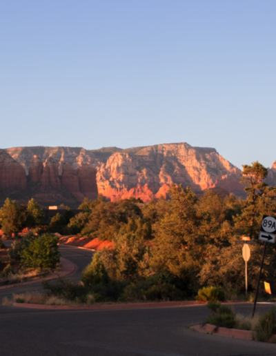 A scenic view of a towering red rock mountain in Sedona with jagged peaks and vibrant orange and red hues, surrounded by desert terrain and clear blue skies.